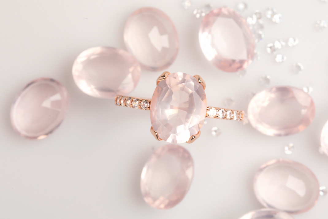 The Rose Quartz Ring 'Harlow' is presented on a white surface and is surrounded by loose Rose Quartz and White Topaz gemstones.