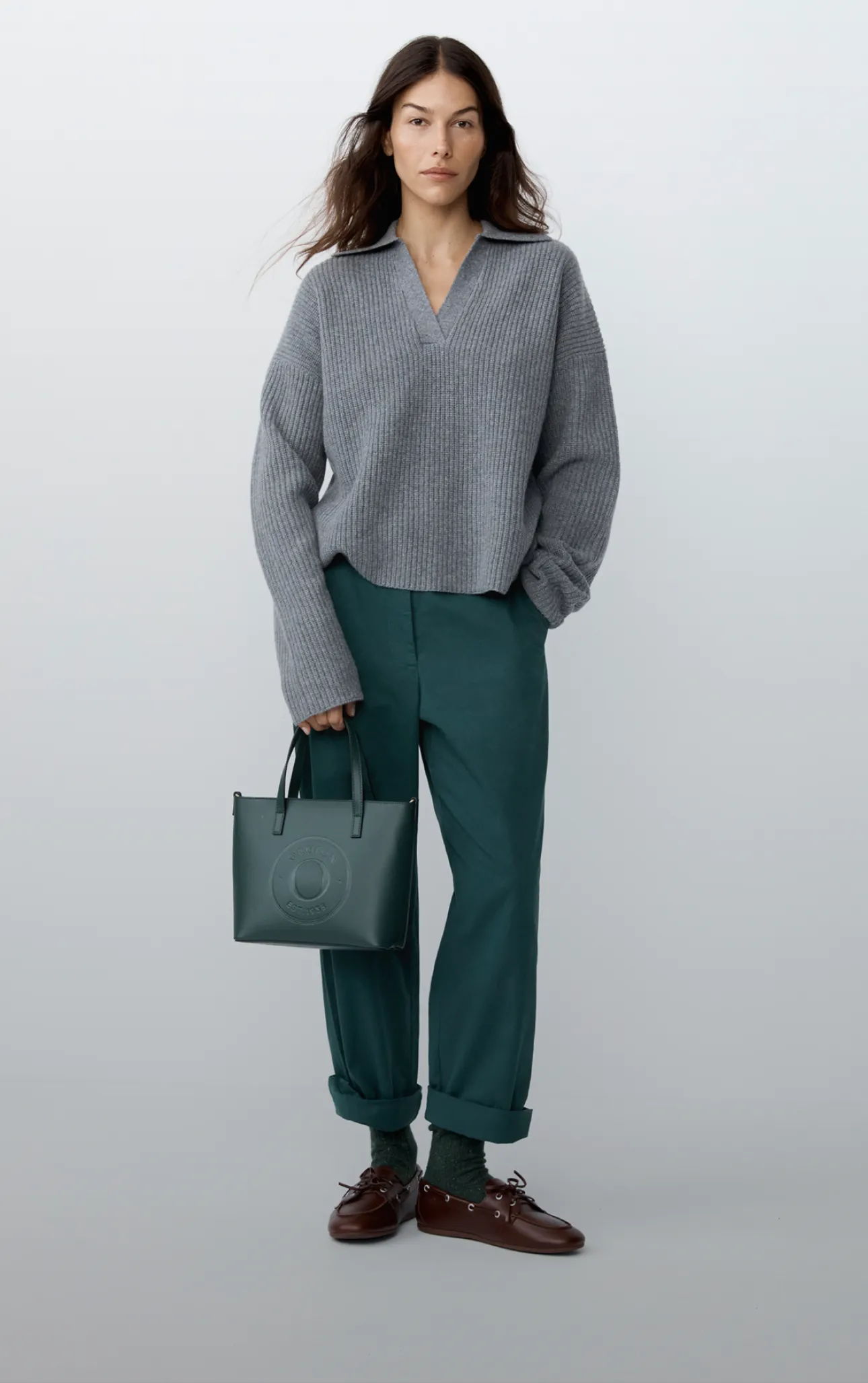 Woman in a gray sweater and green pants holding a handbag.