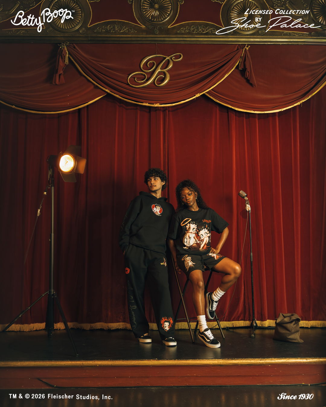 Two individuals posing on stage with red curtains behind them.