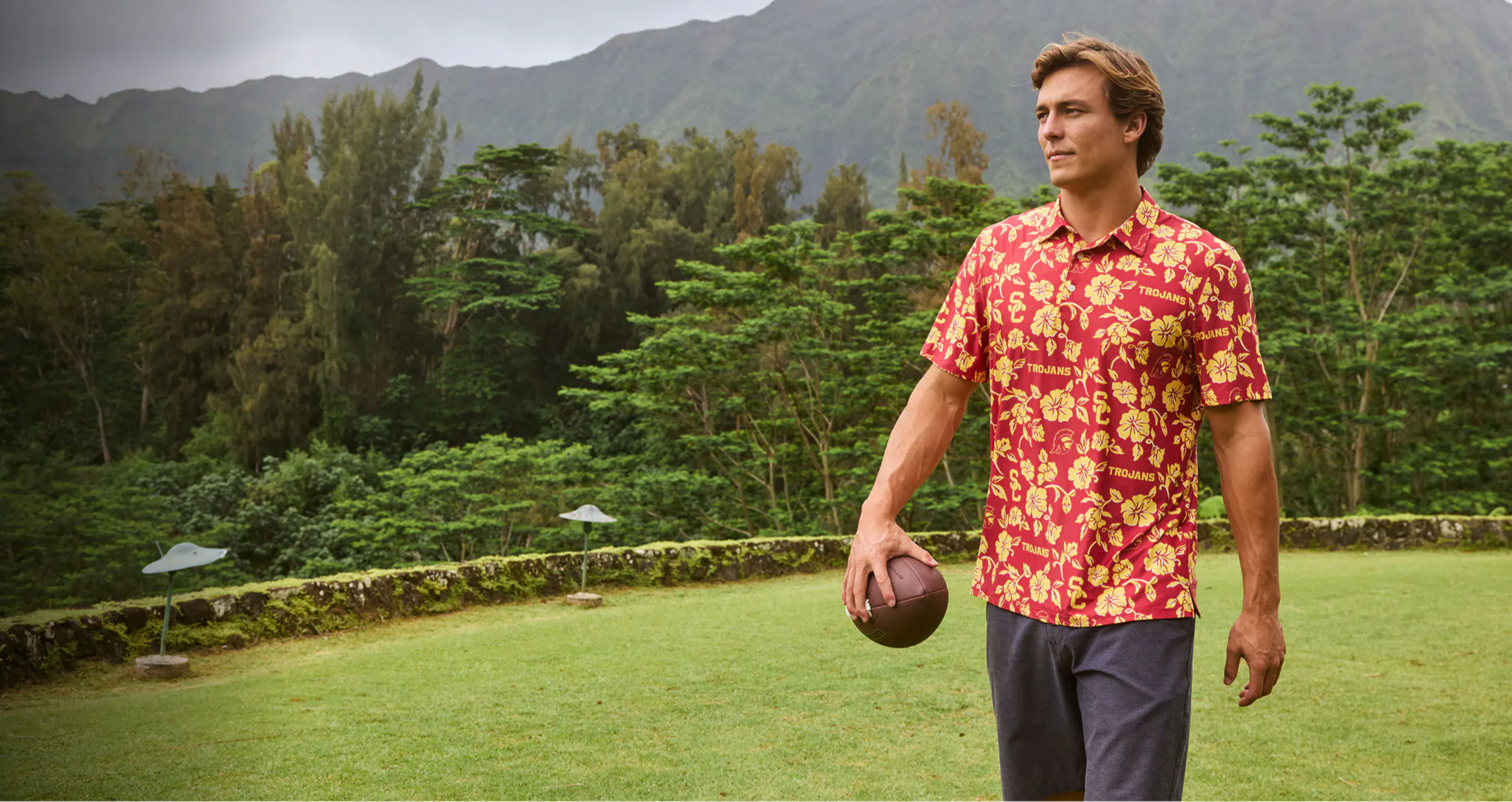 Man in a floral shirt holding a ball in a lush outdoor setting.