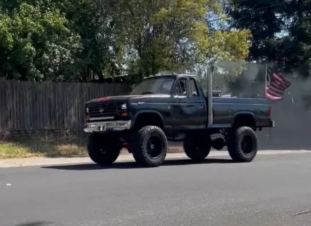 Side view of Trevour's lifted black 1985 Ford F250 with vertical exhaust stack and American flag driving on a paved road