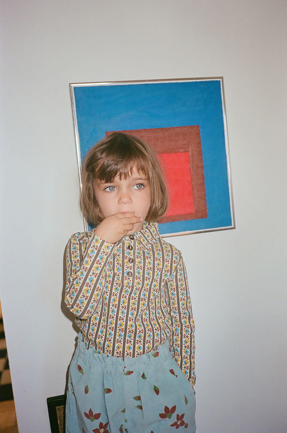 Child with short hair, wearing patterned shirt and blue pants, stands by artwork.