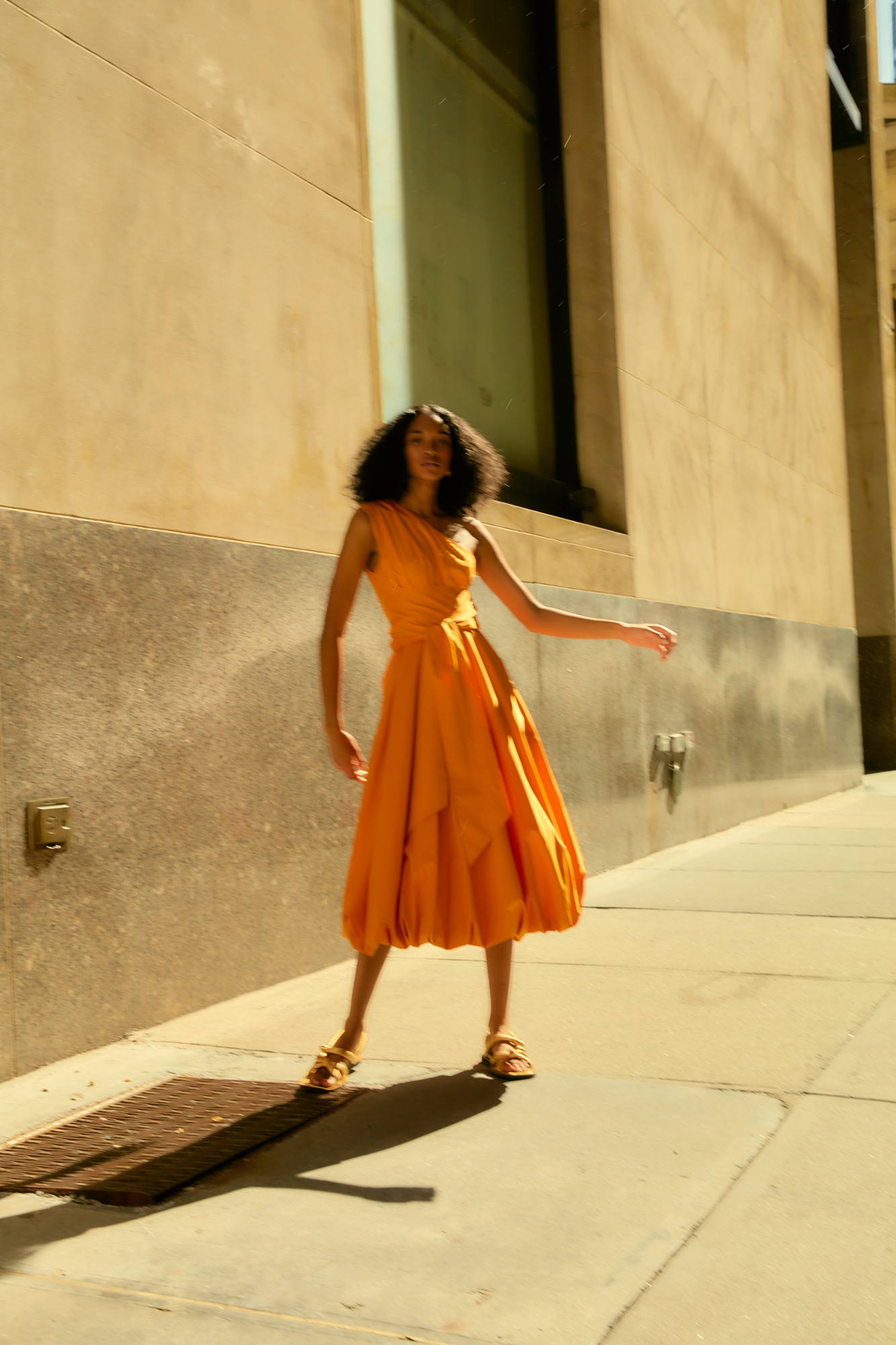 A person in an orange dress poses on a sunny street.