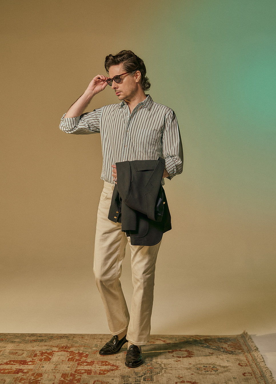 Man in striped shirt and beige pants, holding a jacket, posing stylishly.