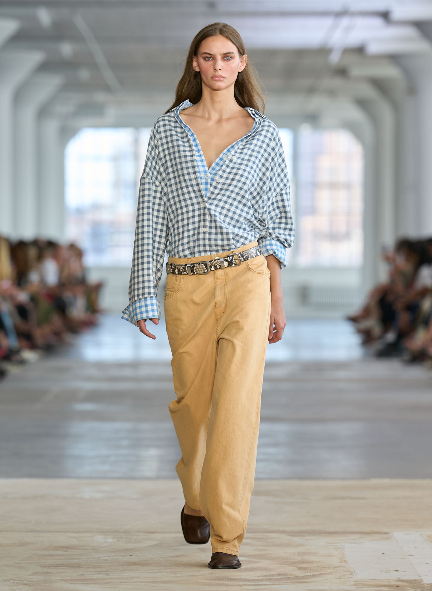 Model walking on runway in casual outfit with checkered shirt and beige pants.