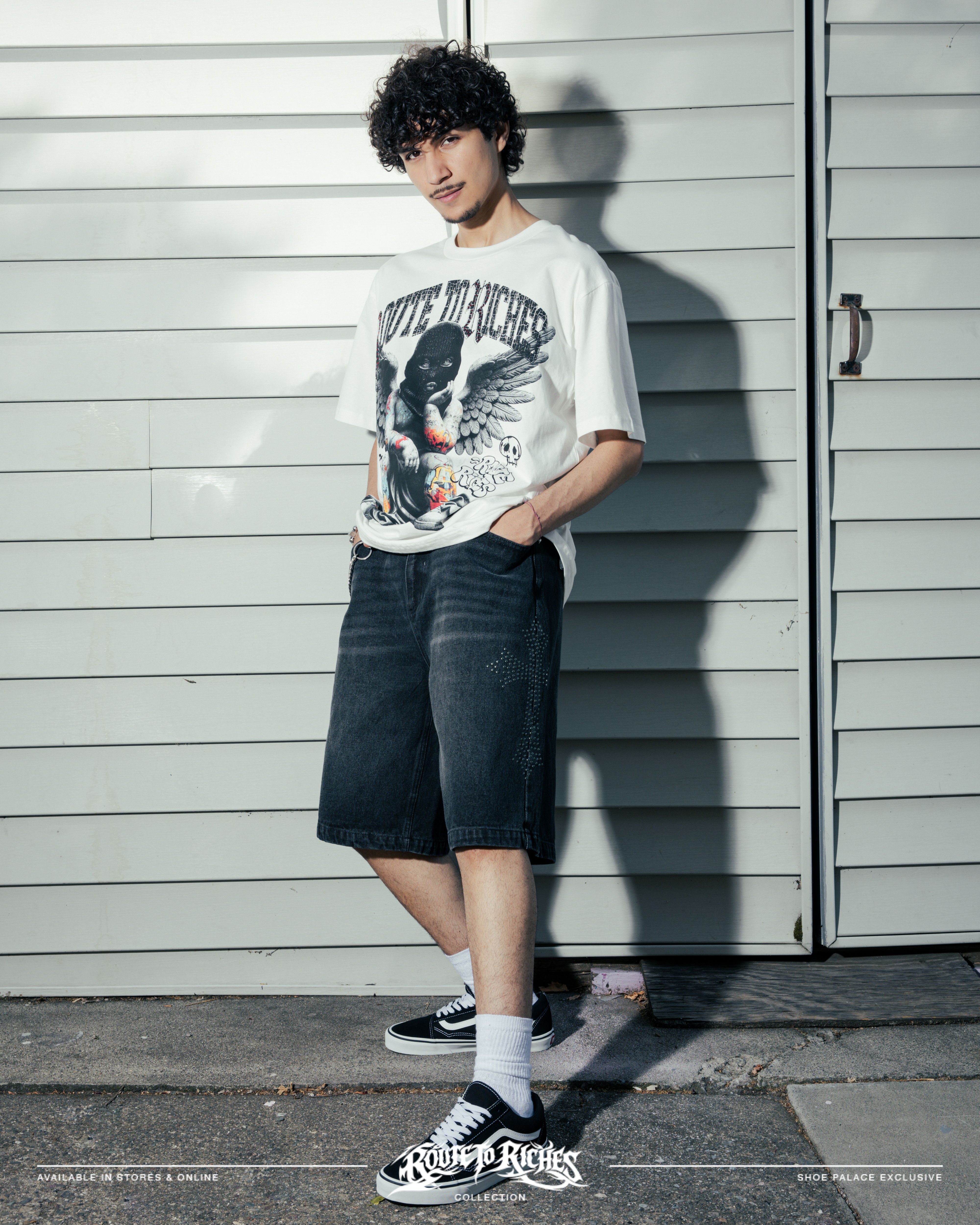 Person in graphic tee and shorts standing against a wall.