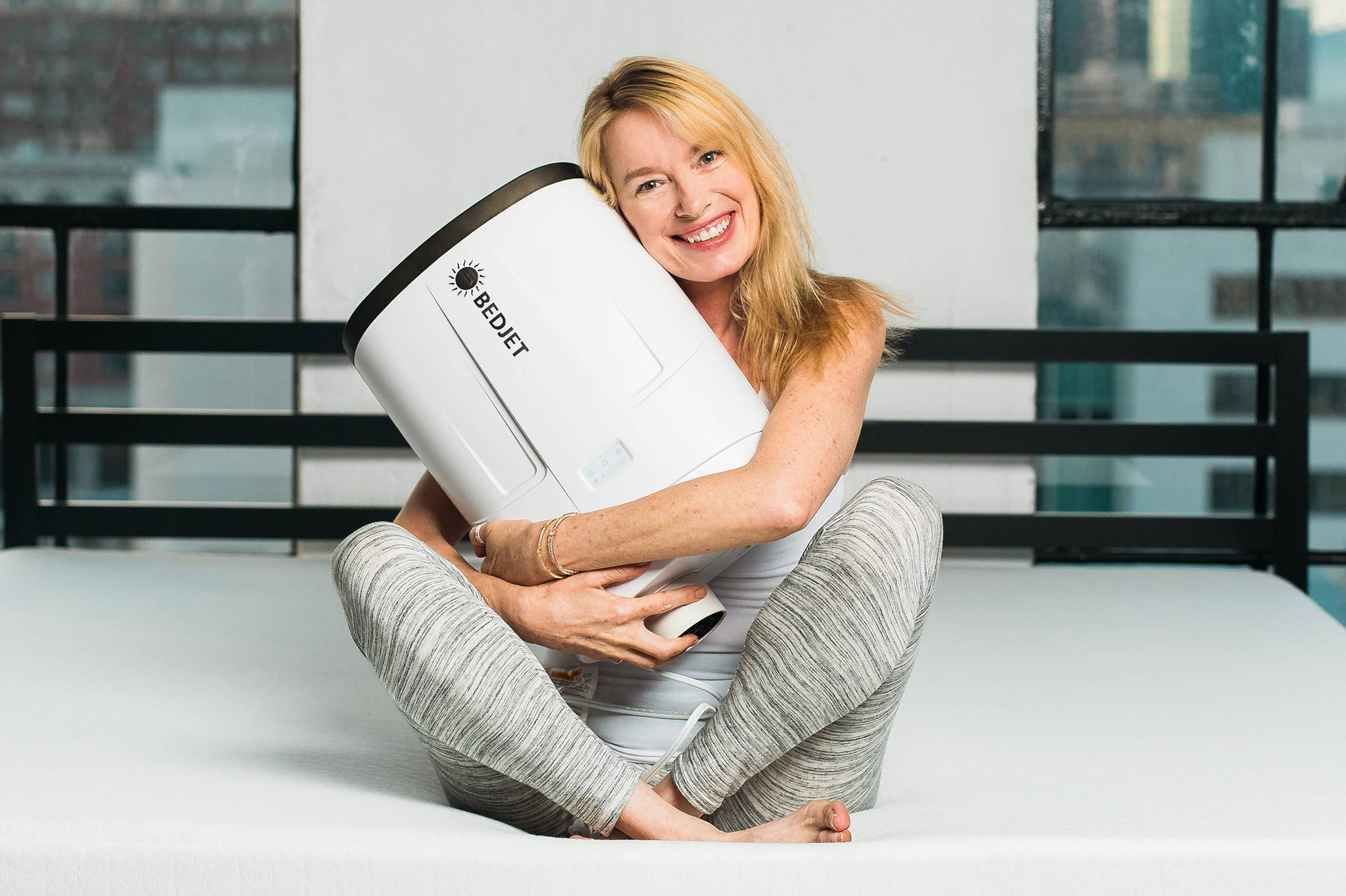 An older blonde woman in gray leggings and a white tank top sitting cross-legged on a bed, holding a BedJet V2 unit in her arms and smiling