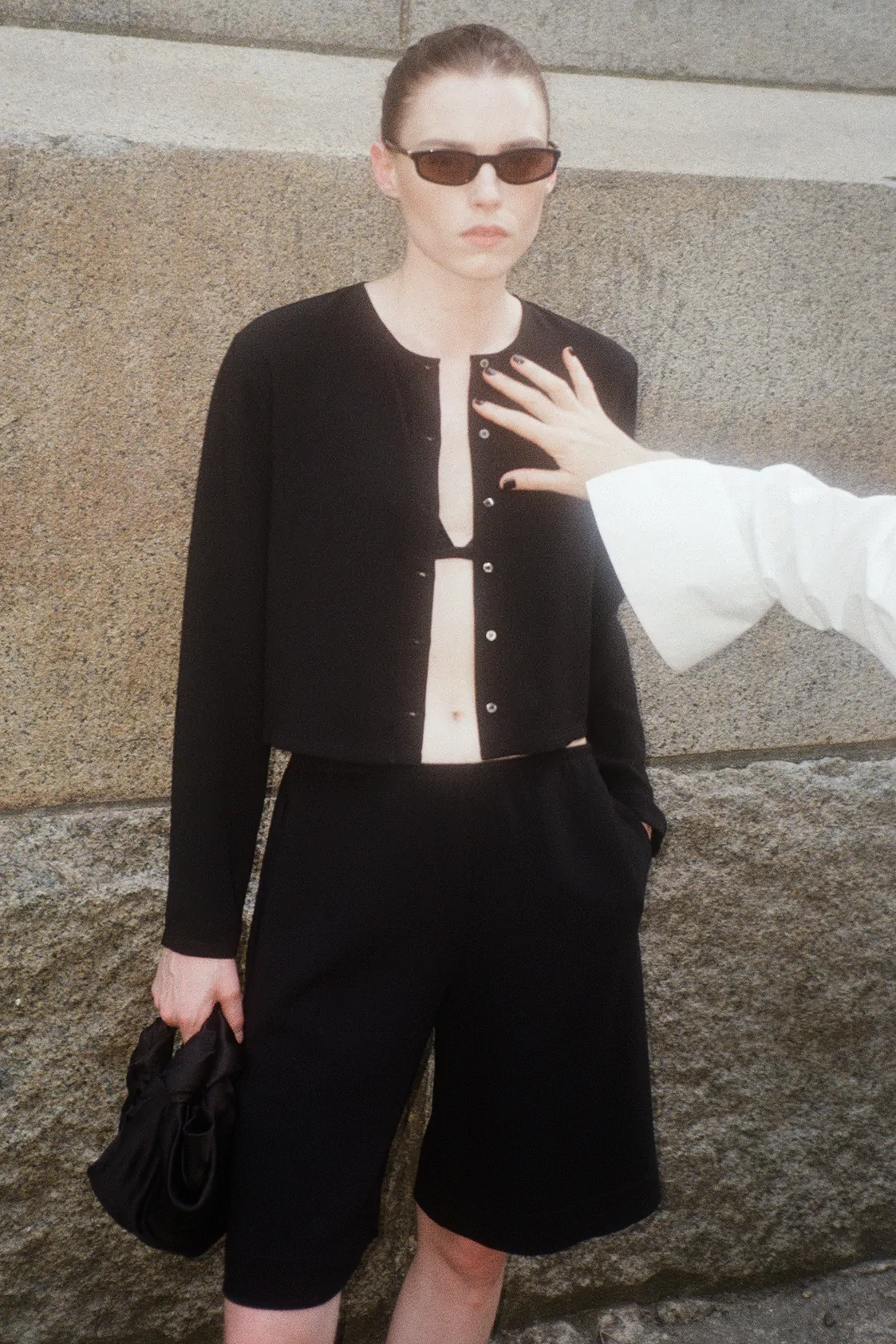 Woman with all black with sunglasses and bag in hand with arm reaching to adjust shirt