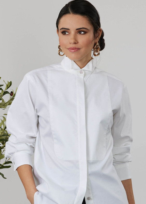 A woman in a white shirt with earrings, standing near greenery.