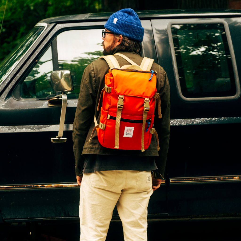 Person wearing a blue beanie and orange backpack near a black vehicle.
