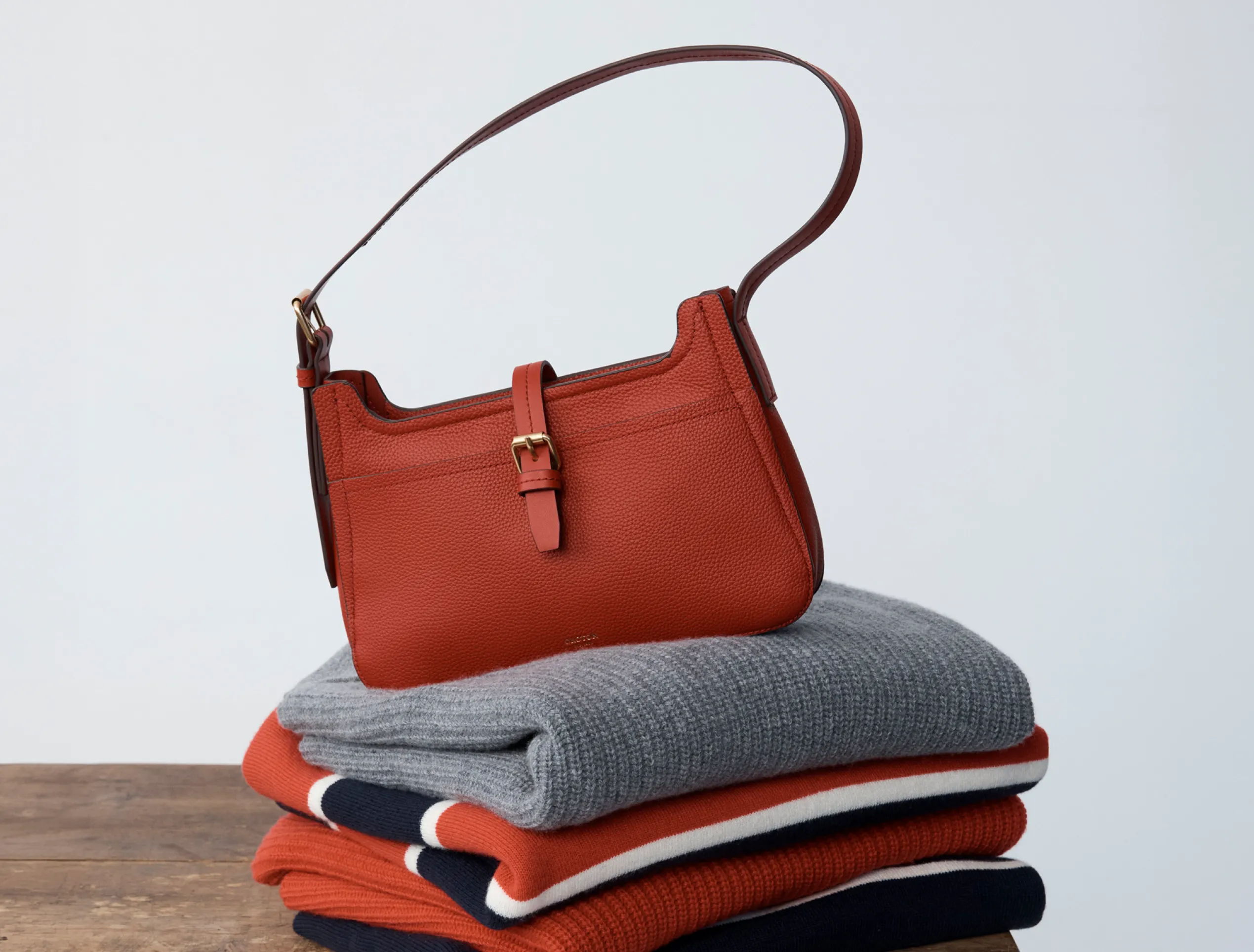 A red handbag on top of folded sweaters.