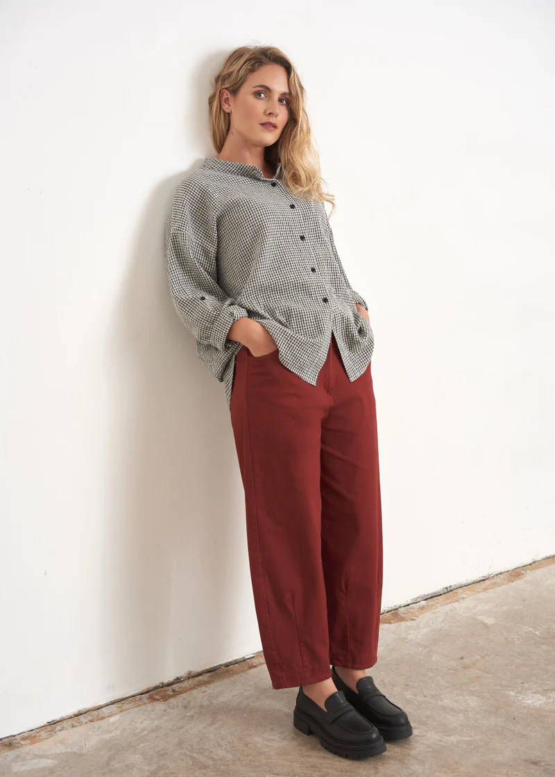 A model wearing a pair of dark red barrel leg trousers with a grey and white checked shirt and black leather loafers