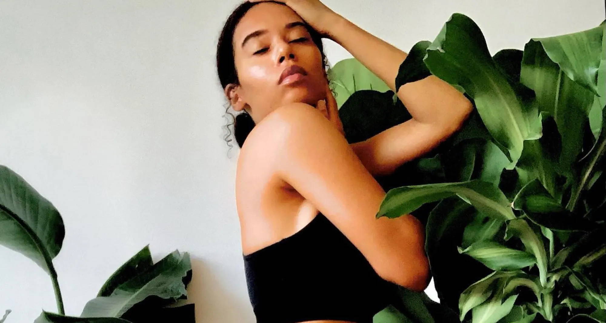 A woman wearing a black bra and underwear touches her own face as she stands in front of a white wall and leafy plants