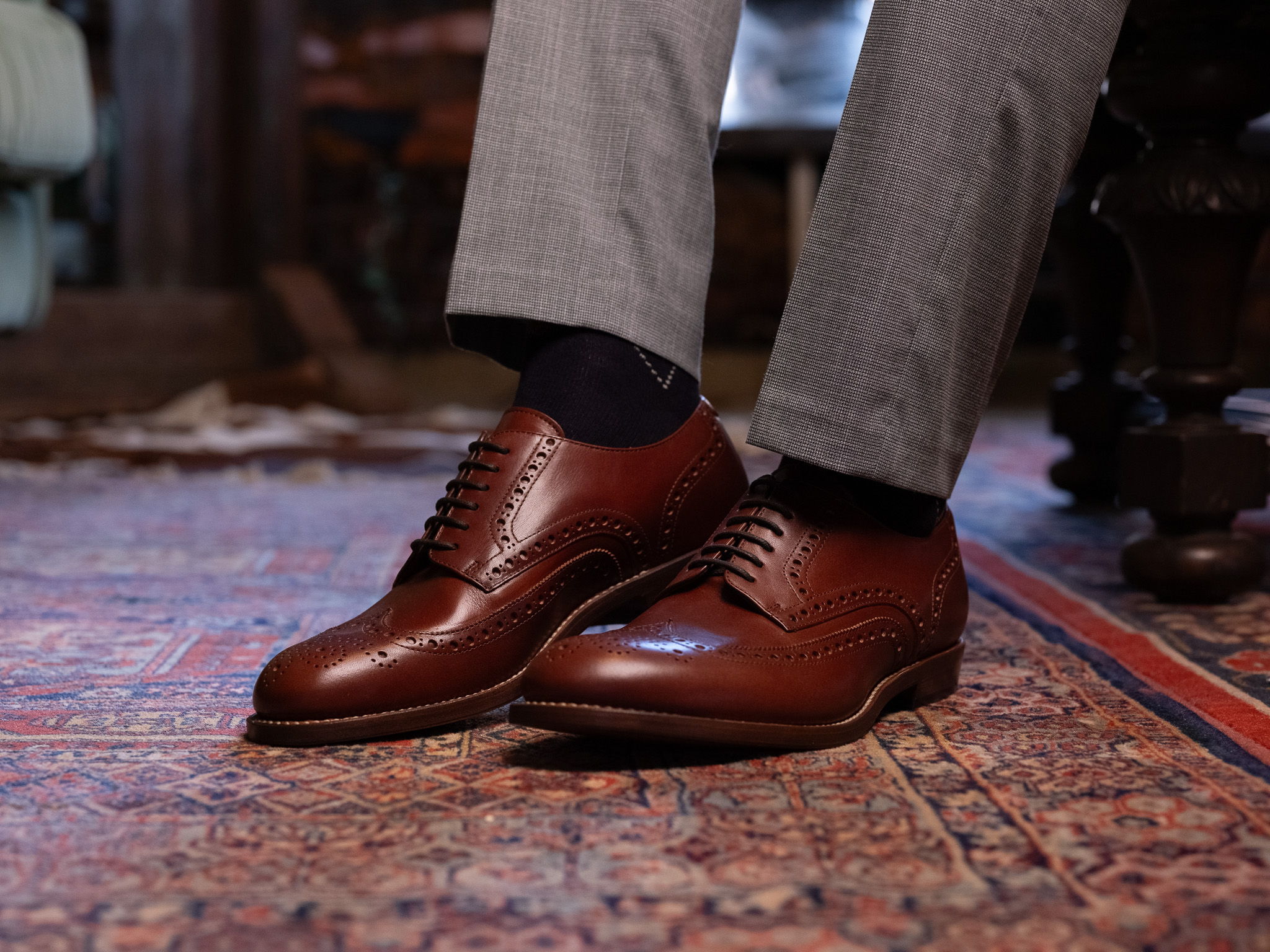 Brown dress shoes worn with gray pants on a patterned rug.