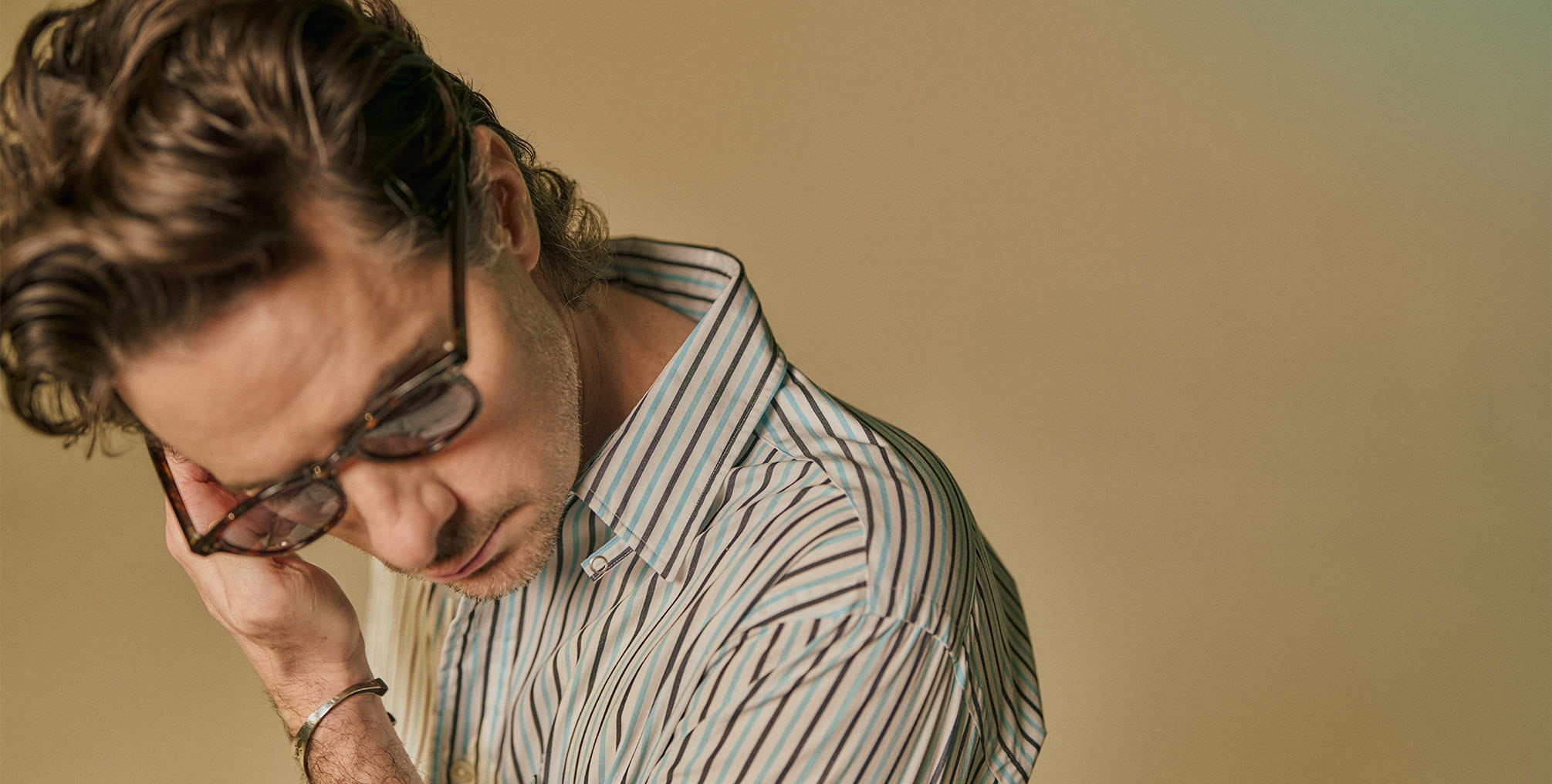 Person with curly hair wearing glasses, looking down against a beige background.
