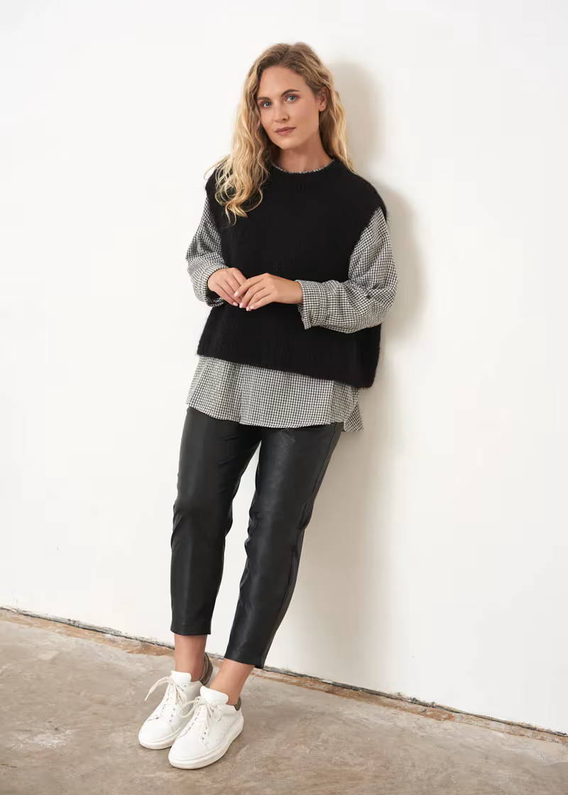 A model wearing a boxy, sleeveless black knitted sleeveless sweater over a black and white checked shirt, black faux leather trousers and white trainers