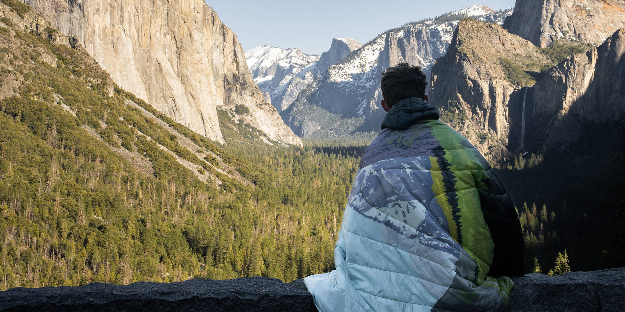 A man sits on a stone wall, wrapped in a Rumpl Original Puffy Blanket, the view of Yosemite National Park can be seen on his blanket and in the background.