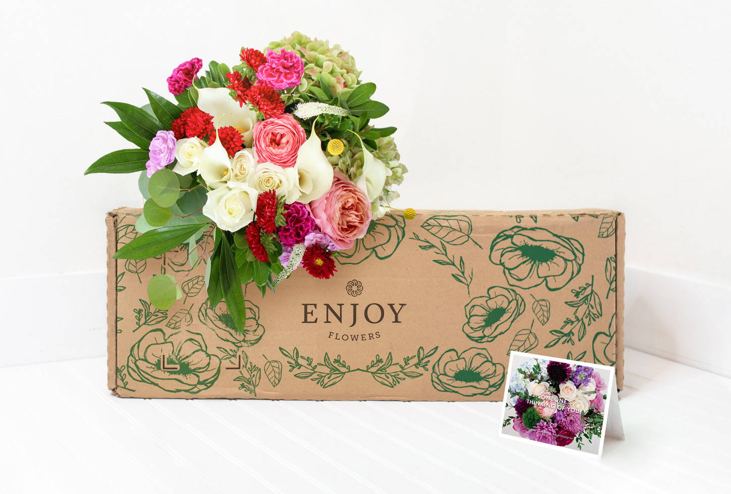 A bright bouquet of premium spring flowers from the Enjoy Flowers Designer Collection sits by a box and personalized card. 