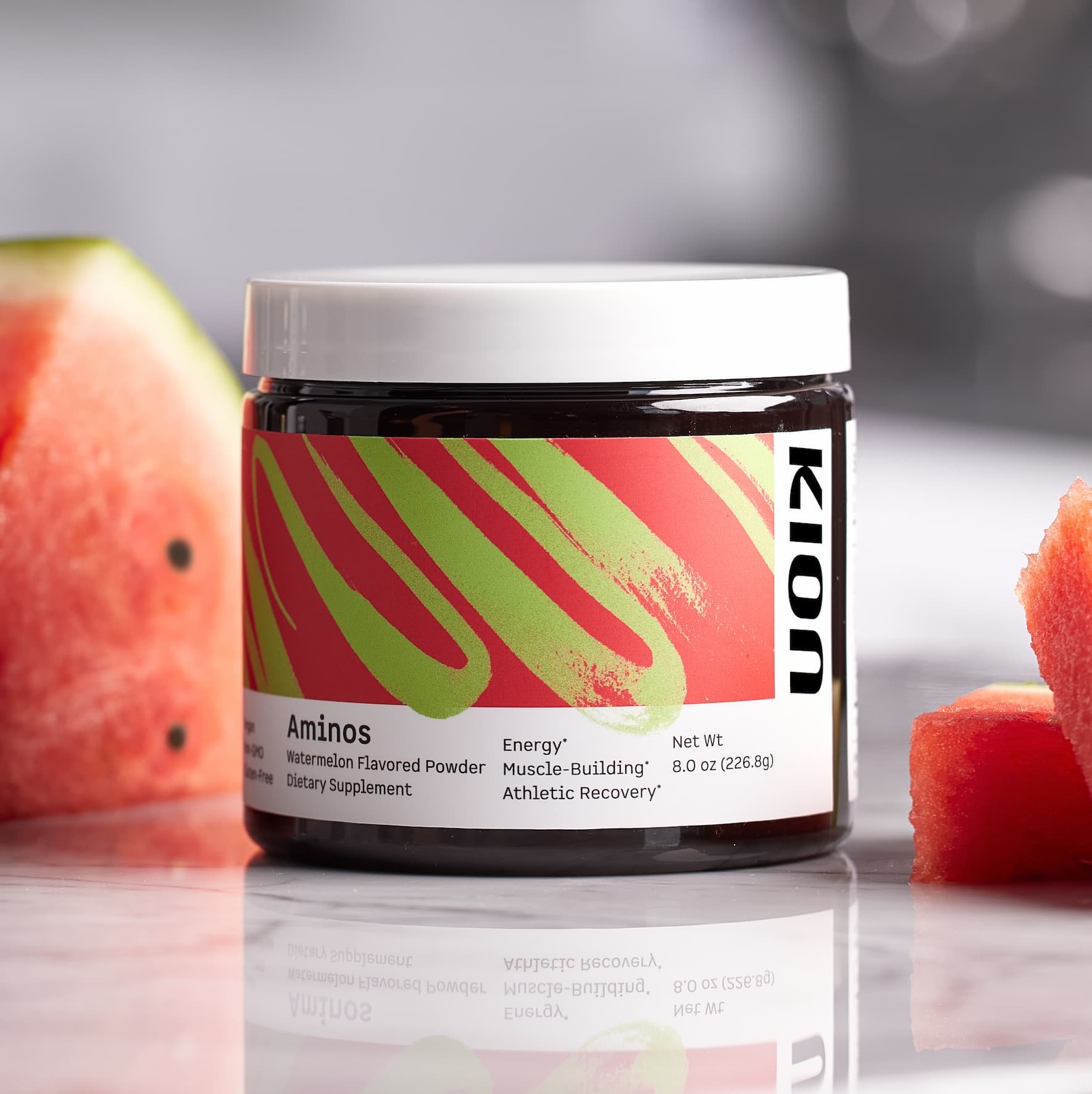 Jar of Kion Aminos Watermelon Flavored Powder dietary supplement with a white lid and vibrant red and green label, displayed on a marble countertop between fresh watermelon slices, with a softly blurred kitchen background.