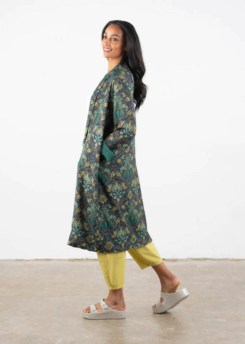 A model wearing a long, green and blue open robe gown with botanical leafy design over yellow trousers and off white chunky platform slides