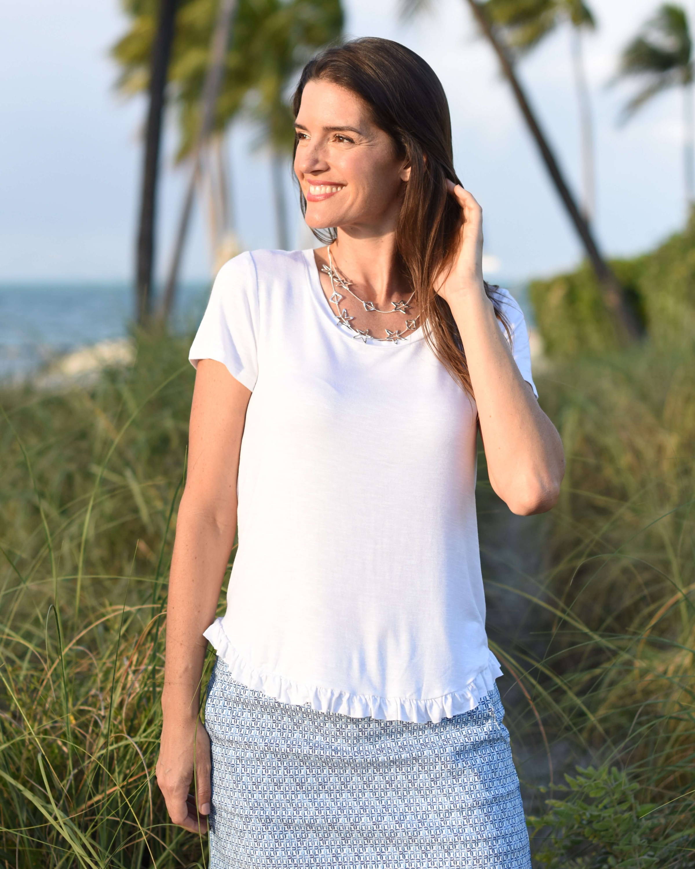 A smiling brunette woman in a white top and blue skort runs her hand through her hair at the beach. 