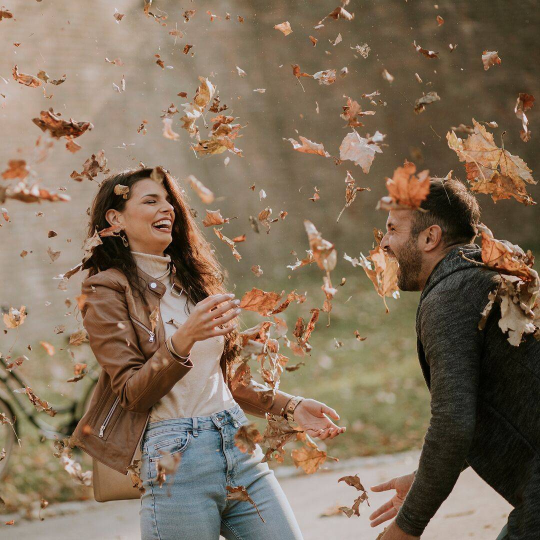 Couple laughing at the joyful whirl of fall leaves tossed up in the air by a gust of wind