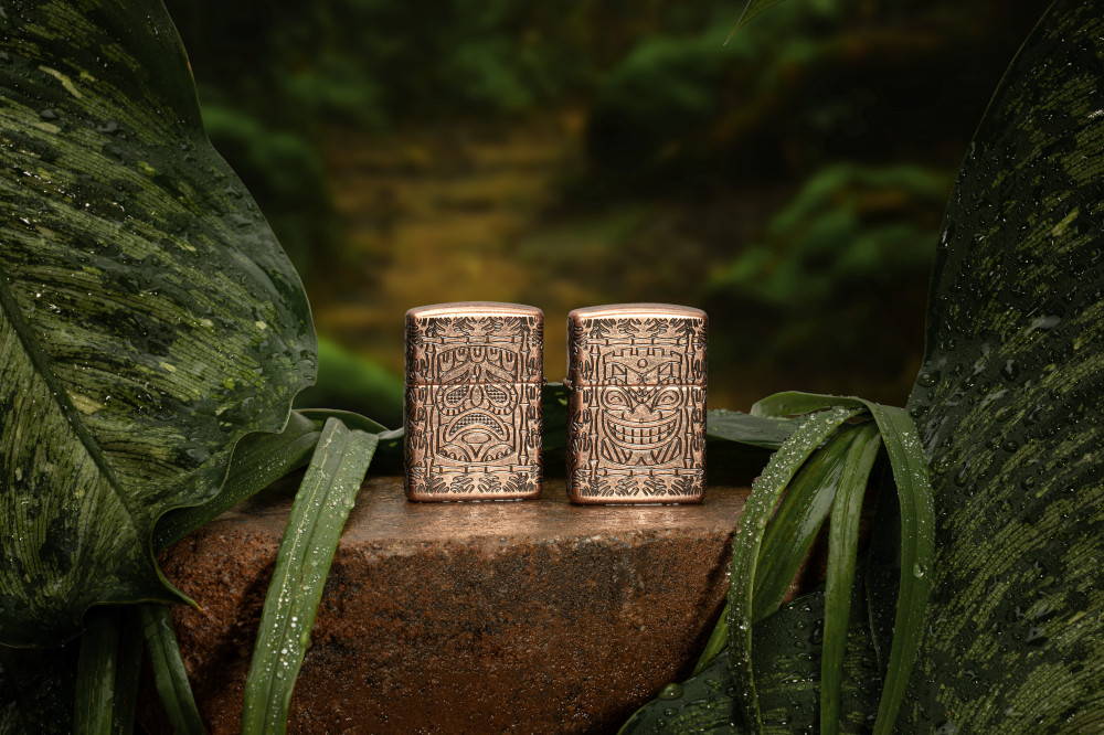 Lifestyle image of two Tiki Design Armor® Antique Copper Windproof Lighters, one showing the front of the design and the other showing the back. The lighters are standing on a stone in a jungle theme.