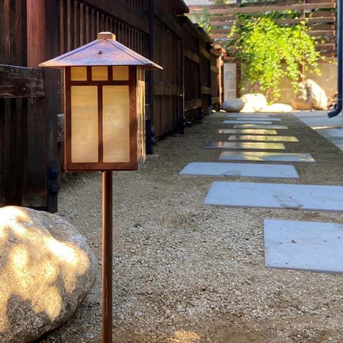 A Craftsman style pathway lantern with a raw copper finish next to sand and pavers.