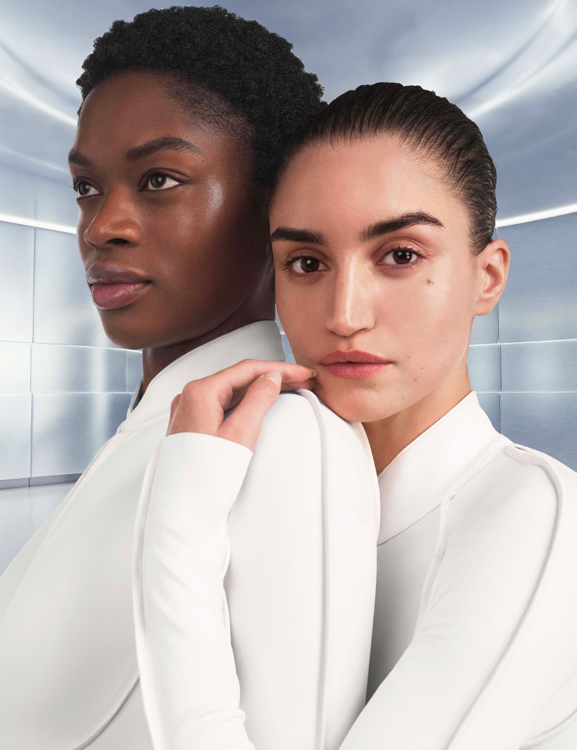 Two women in sleek white outfits, standing close together, with a futuristic background.