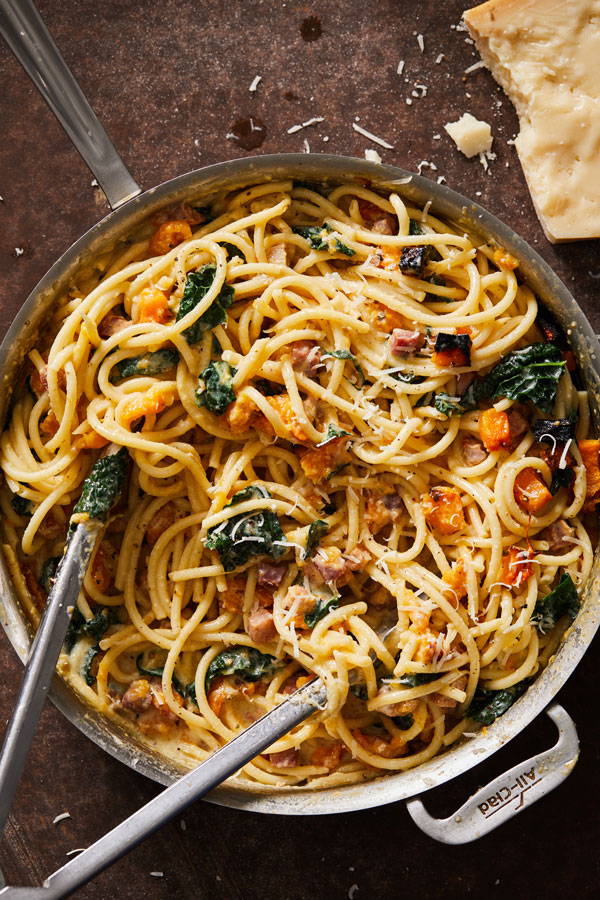 Bucatini pasta in a light creamy sauce tossed with cubed pancetta and butternut squash and prepared in a skillet with tongs for serving