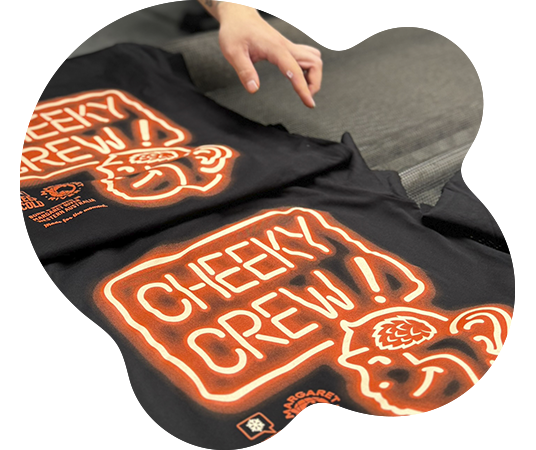 Hands reaching for a folded black t-shirt printed with a bold orange neon-style Cheeky Crew graphic with illustrated monkey character