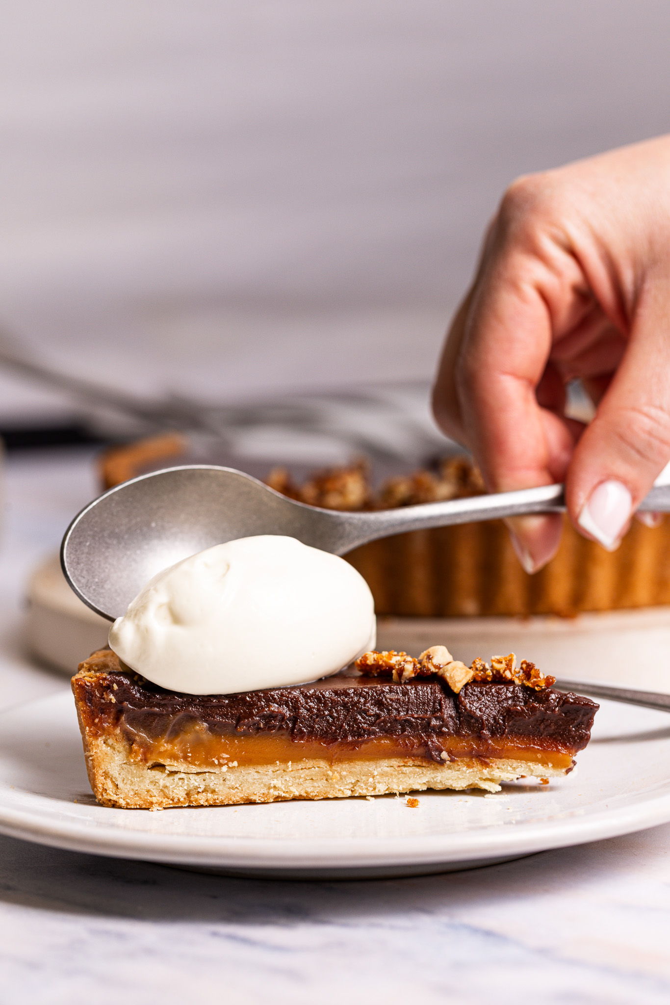 A hand adds whipped cream to a slice of chocolate tart on a plate.