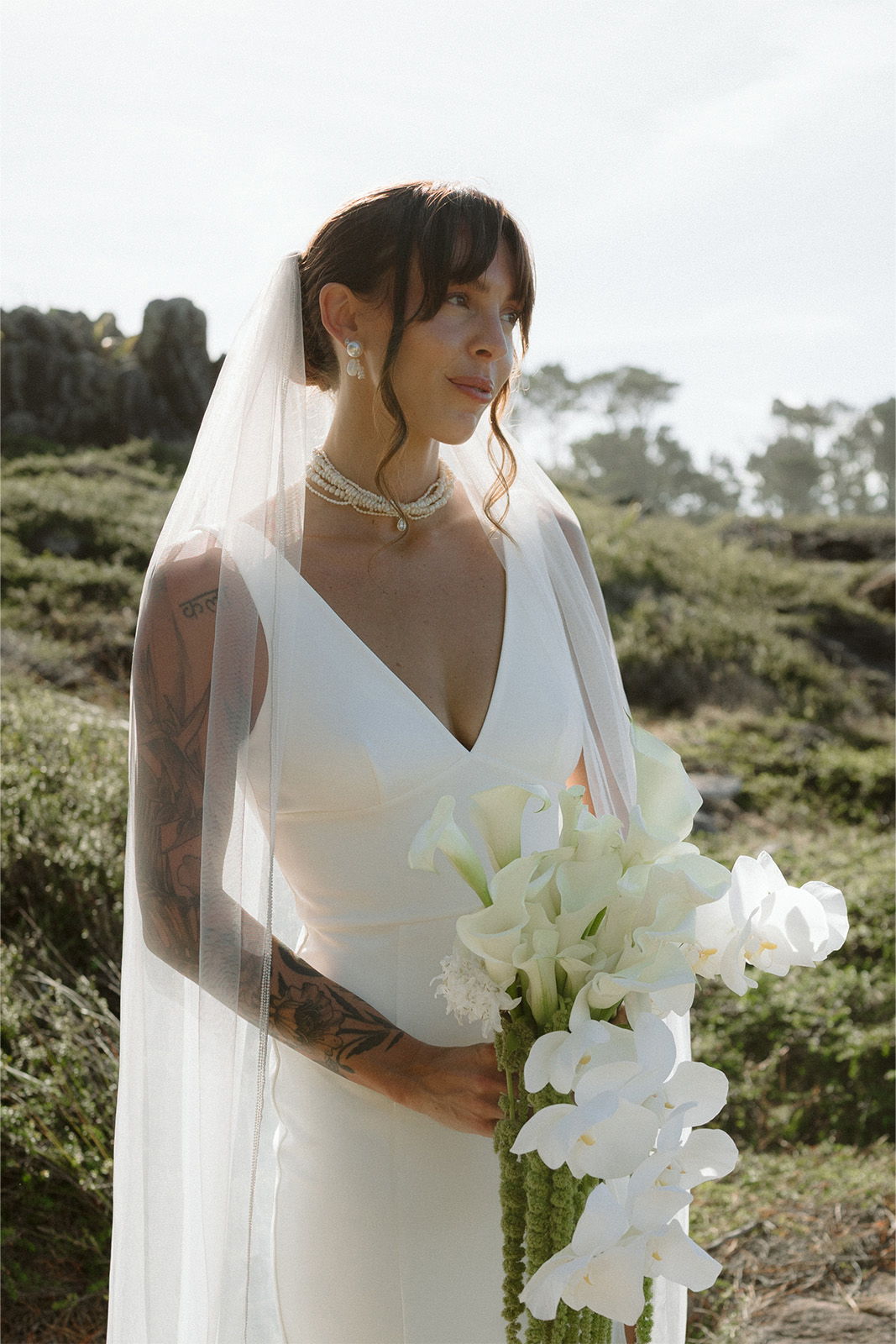 Bride in white dress holding white flowers, with a veil outdoors.