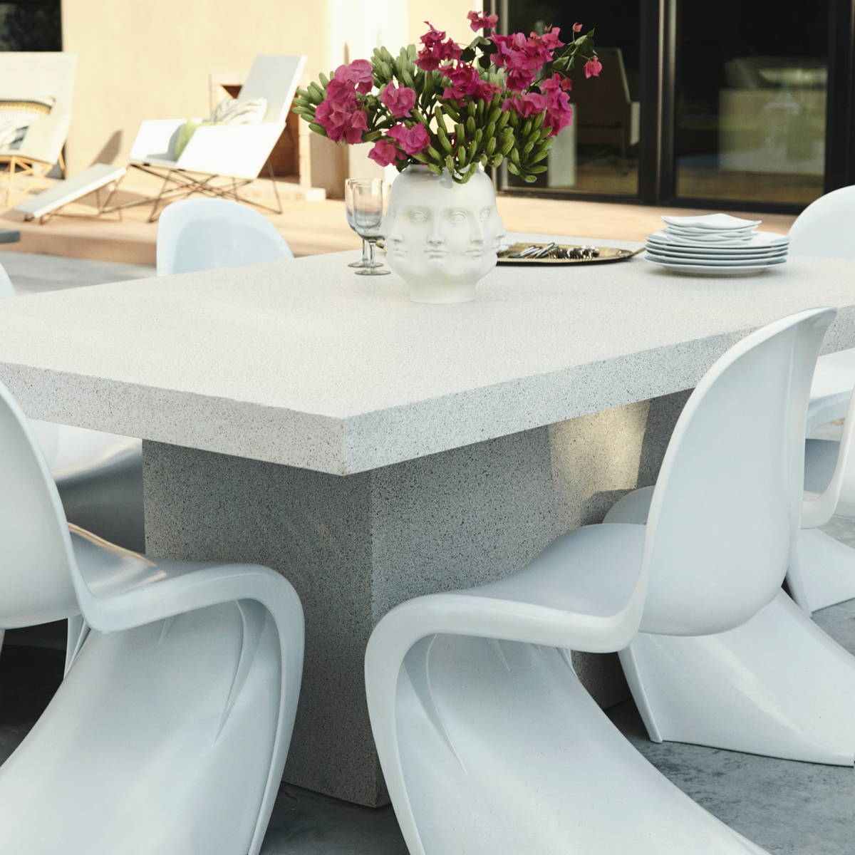 Modern lightweight concrete table with curved white chairs and a centerpiece pot adorned in faces and filled with pink flowers.