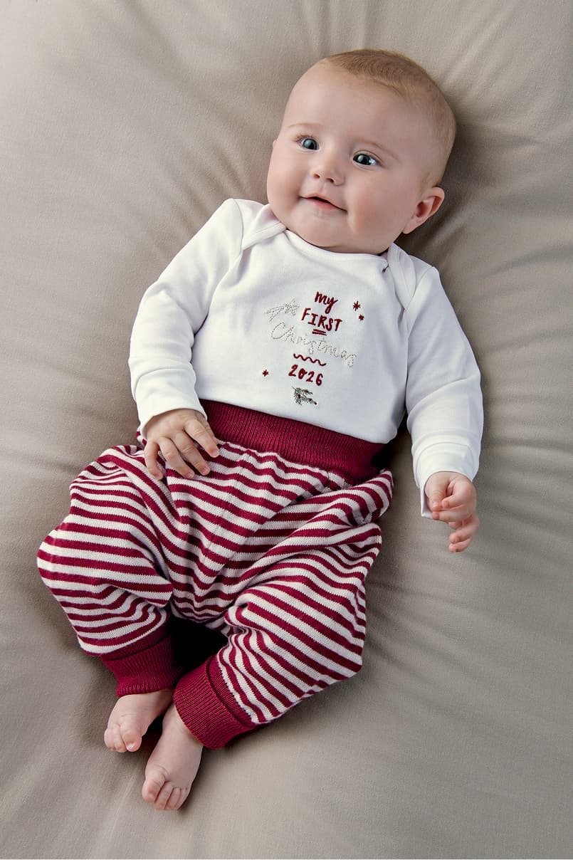 Baby lying on a beige surface, wearing a white shirt and striped pants.