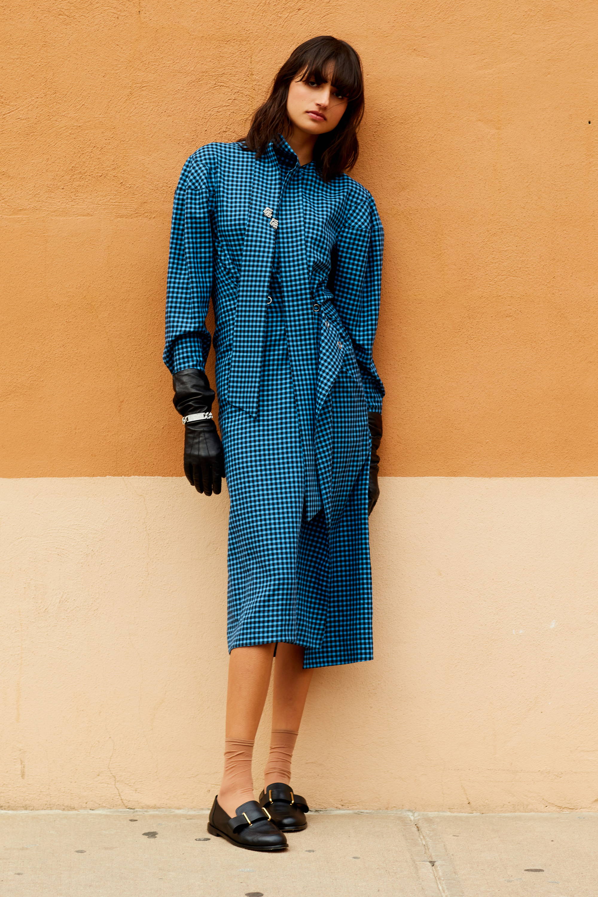 Model wearing a blue patterned dress with gloves against a beige wall.