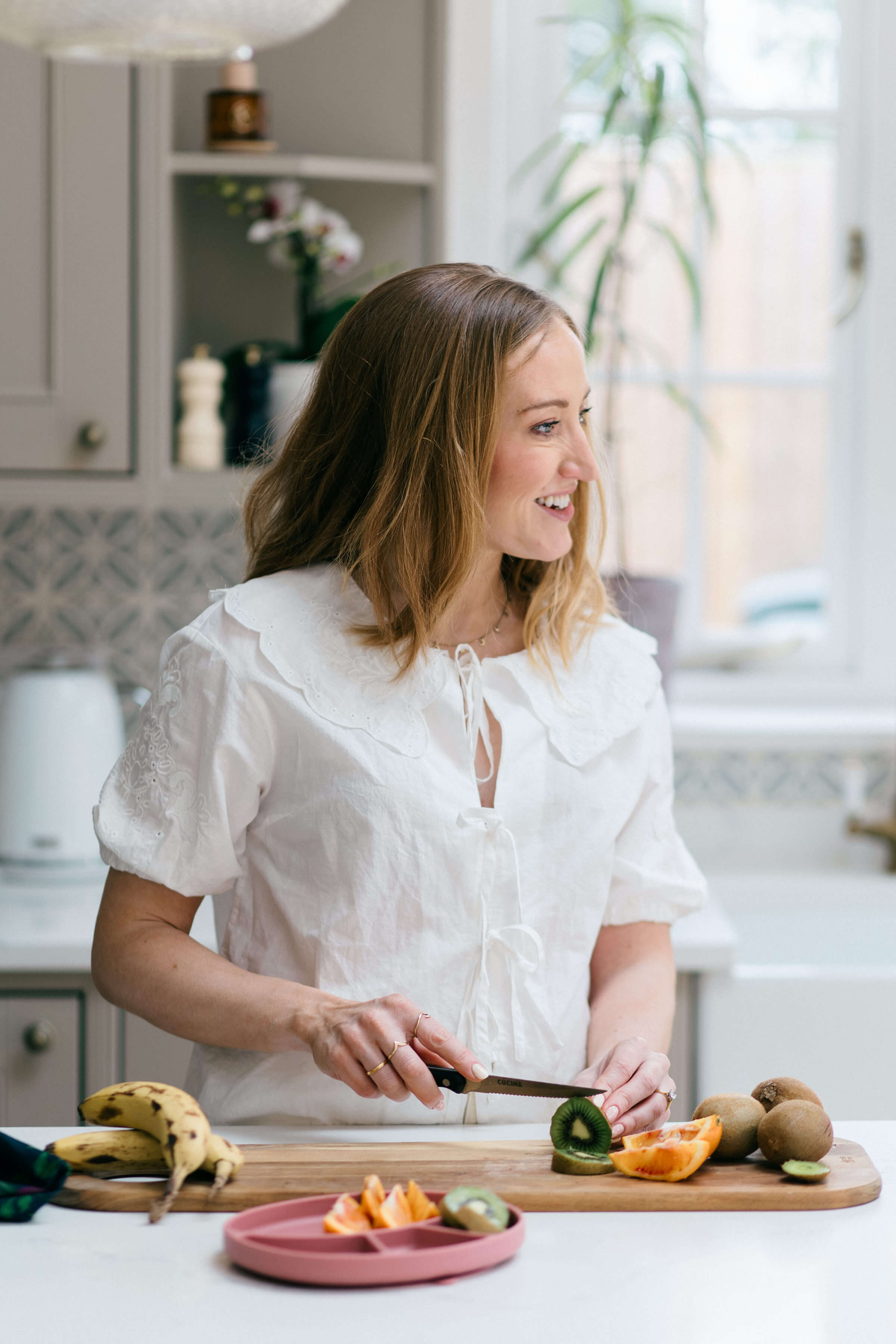 Woman chopping vegetables in a bright kitchen, smiling with fruits on the counter.