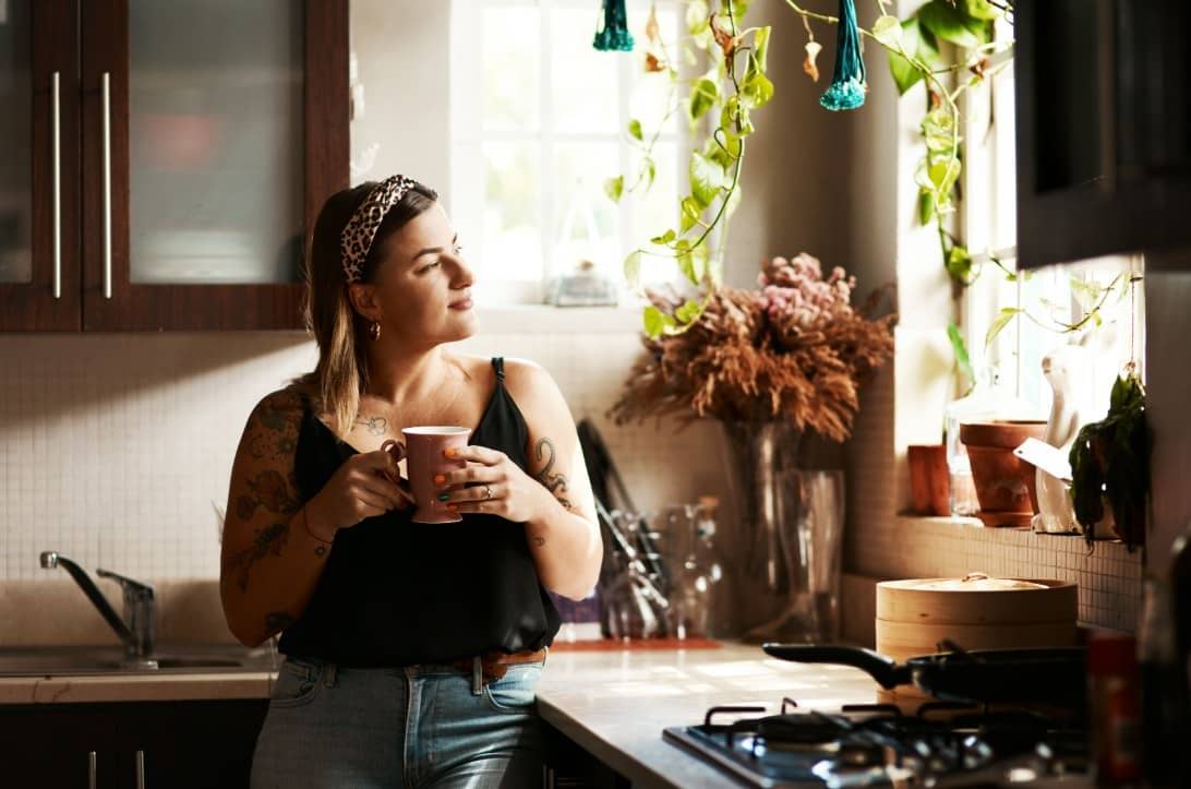 A woman leans against a sunlit kitchen counter, holding a mug and gazing out the window