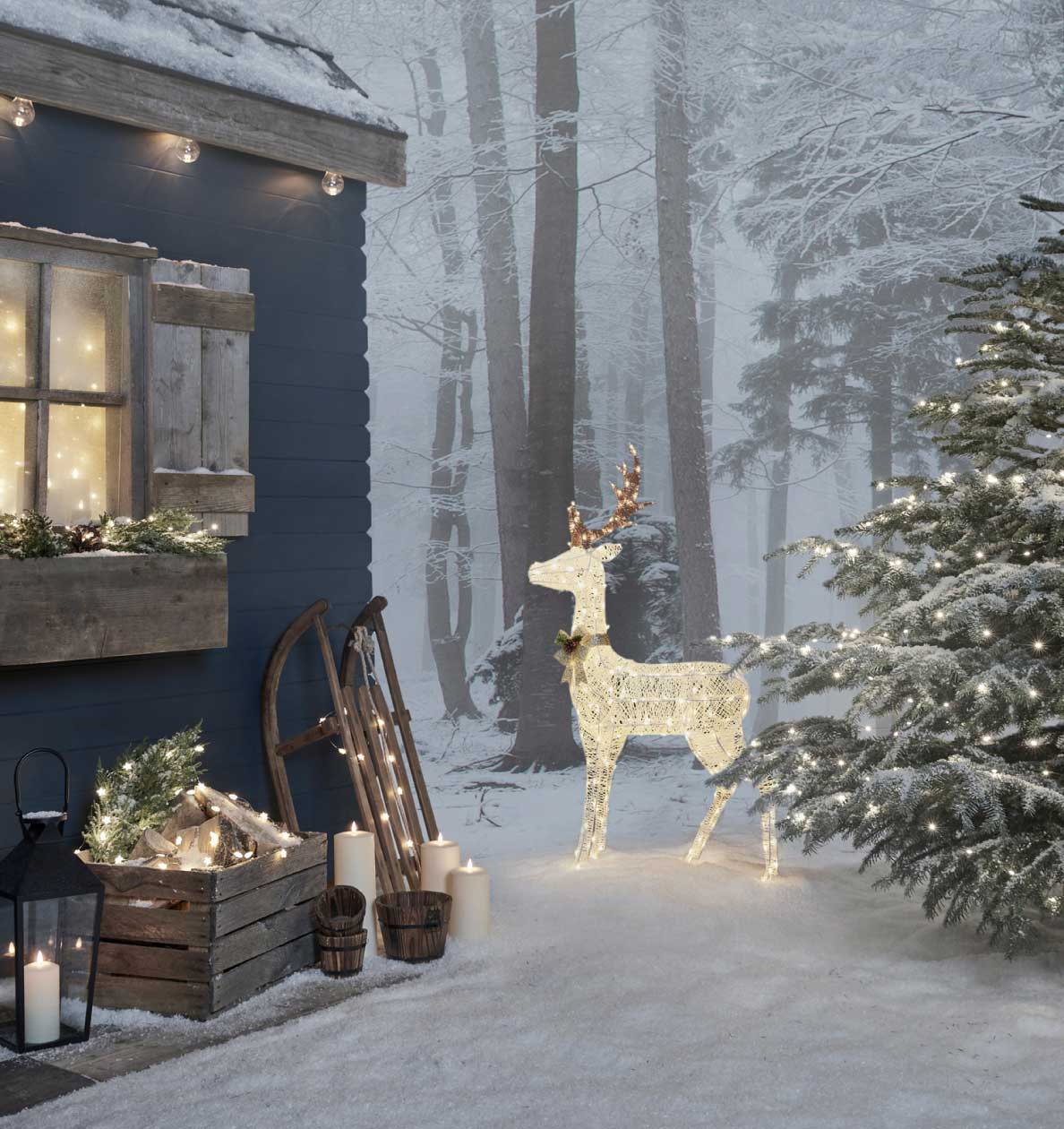 LED Rentier im Schnee zwischen einer beleuchteten Holzhütte und einem beleuchteten Tannenbaum