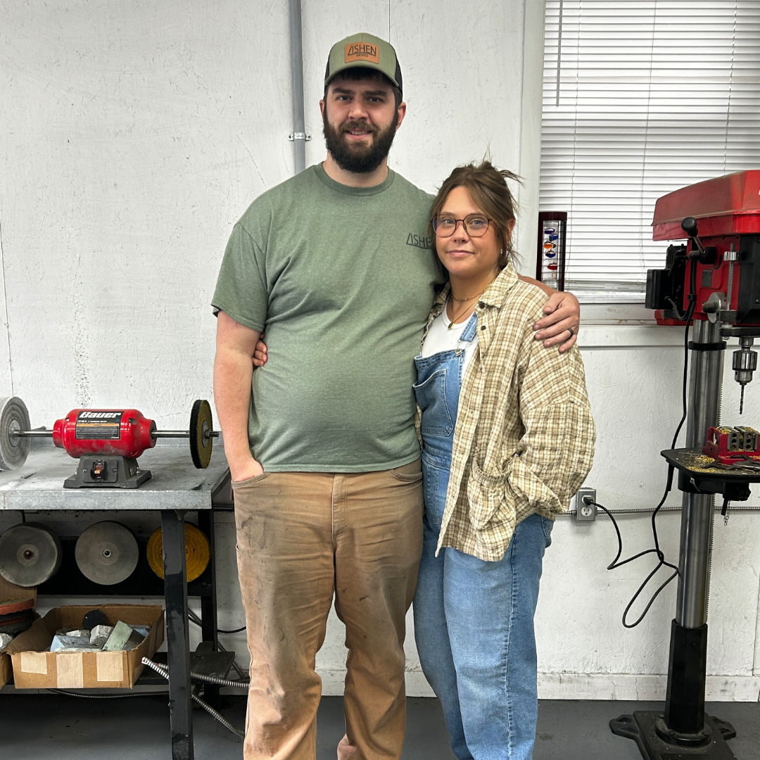 Two people smiling in a workshop with tools and equipment around them.