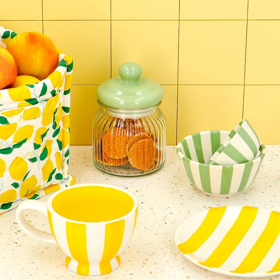 A kitchen counter display showcasing lemon-themed kitchenware including a yellow-striped mug, green and white striped bowls, and a green lid jar, complemented by a fruit-filled lemon patterned fabric bag.