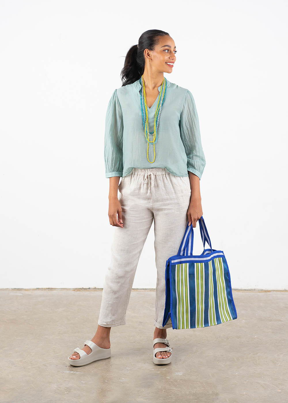 A model wearing an aqua blue lightweight cotton blouse with off white trousers, colourful beaded necklaces, off white chunky platform slides and holding a blue and yellow stripy shopping bag