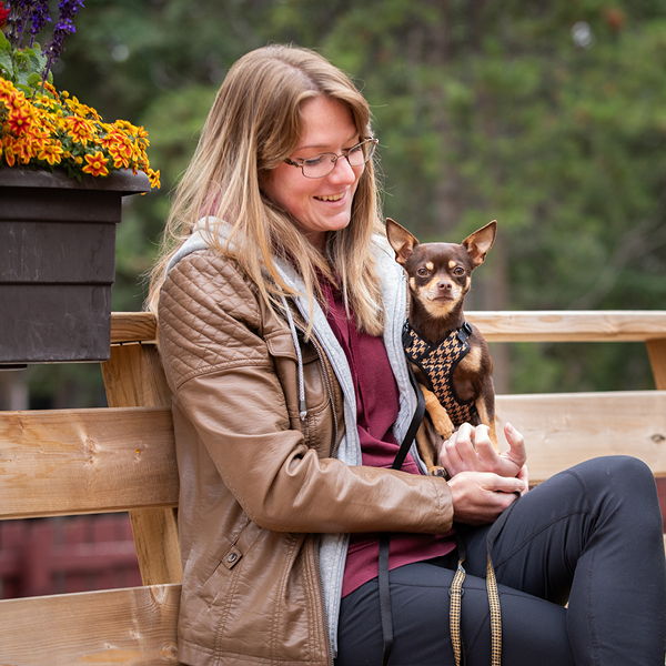 Woman sitting on a bench holding a small dog with flowers in the background.