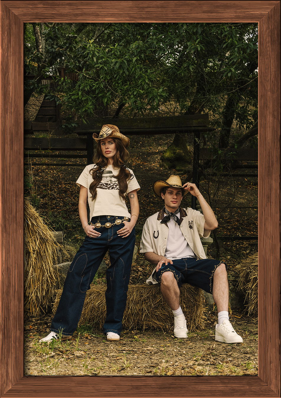 Two individuals in cowboy hats, posing outdoors near hay bales.