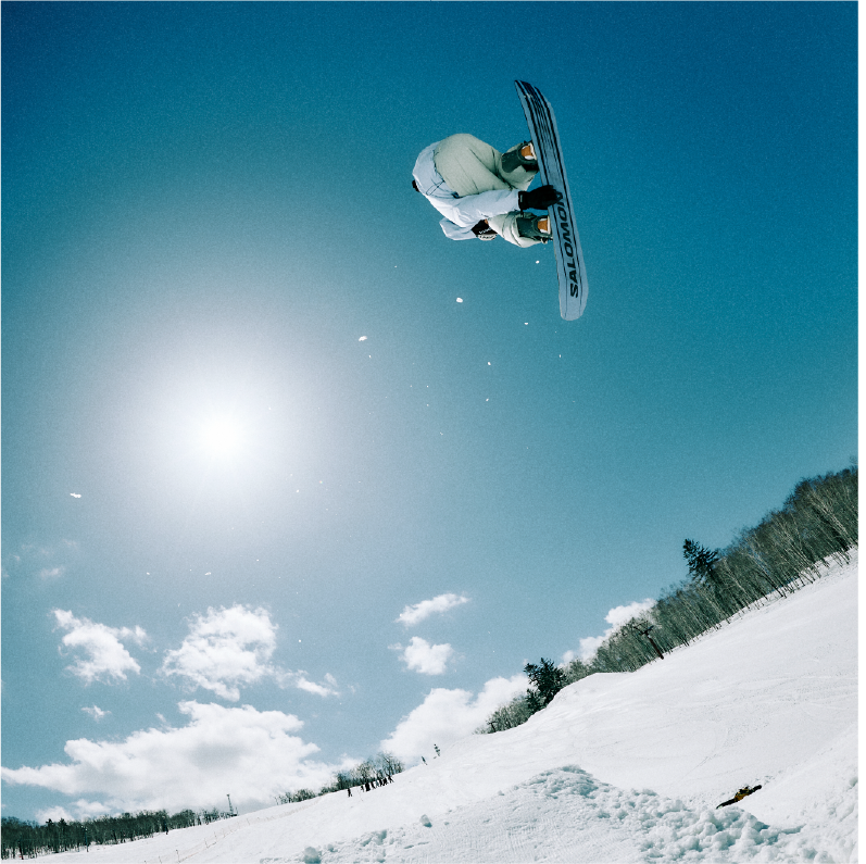 Snowboarder performing a jump against a bright sun and blue sky.