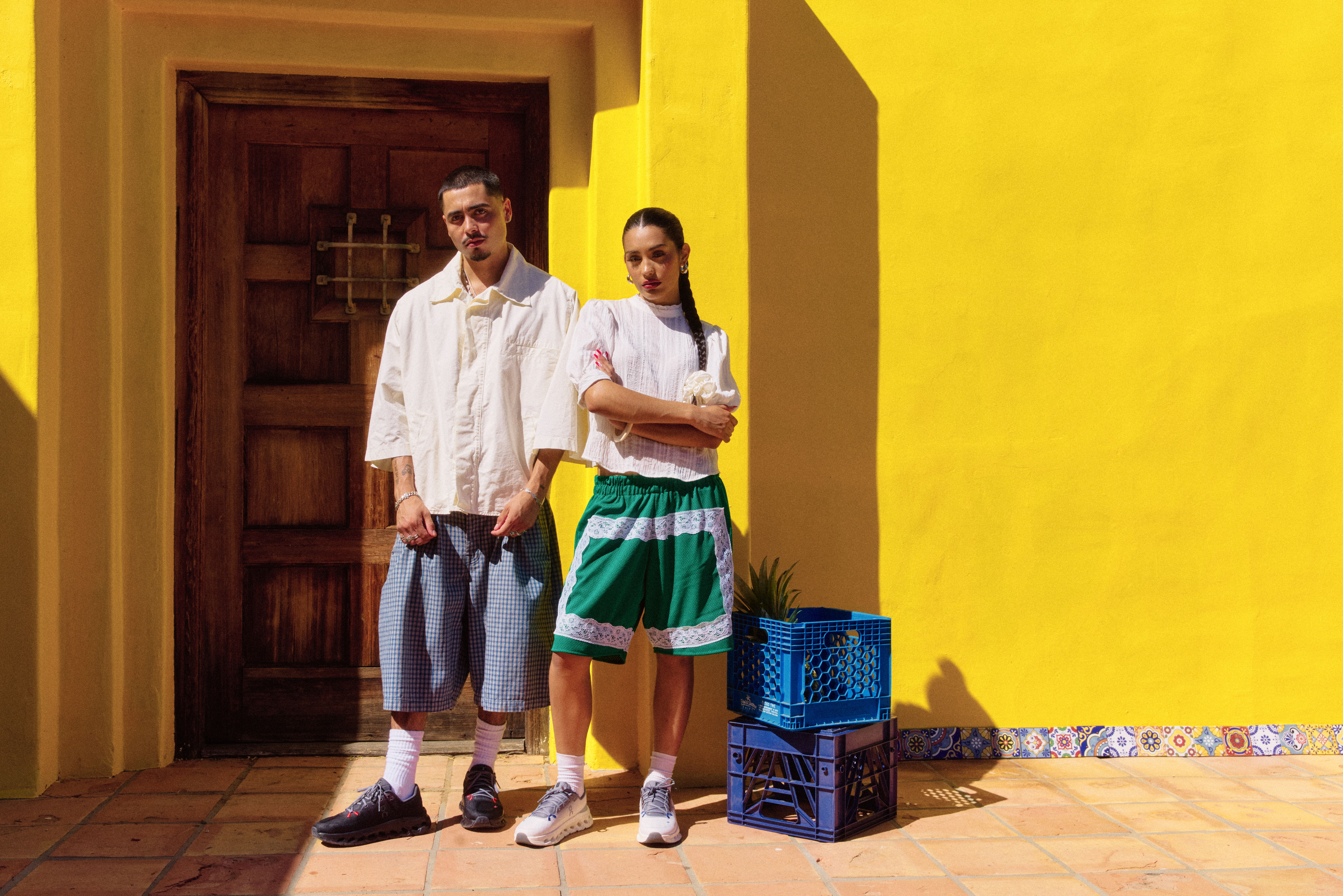 Two people standing against a bright yellow wall, casual outfits, one holding a crate.