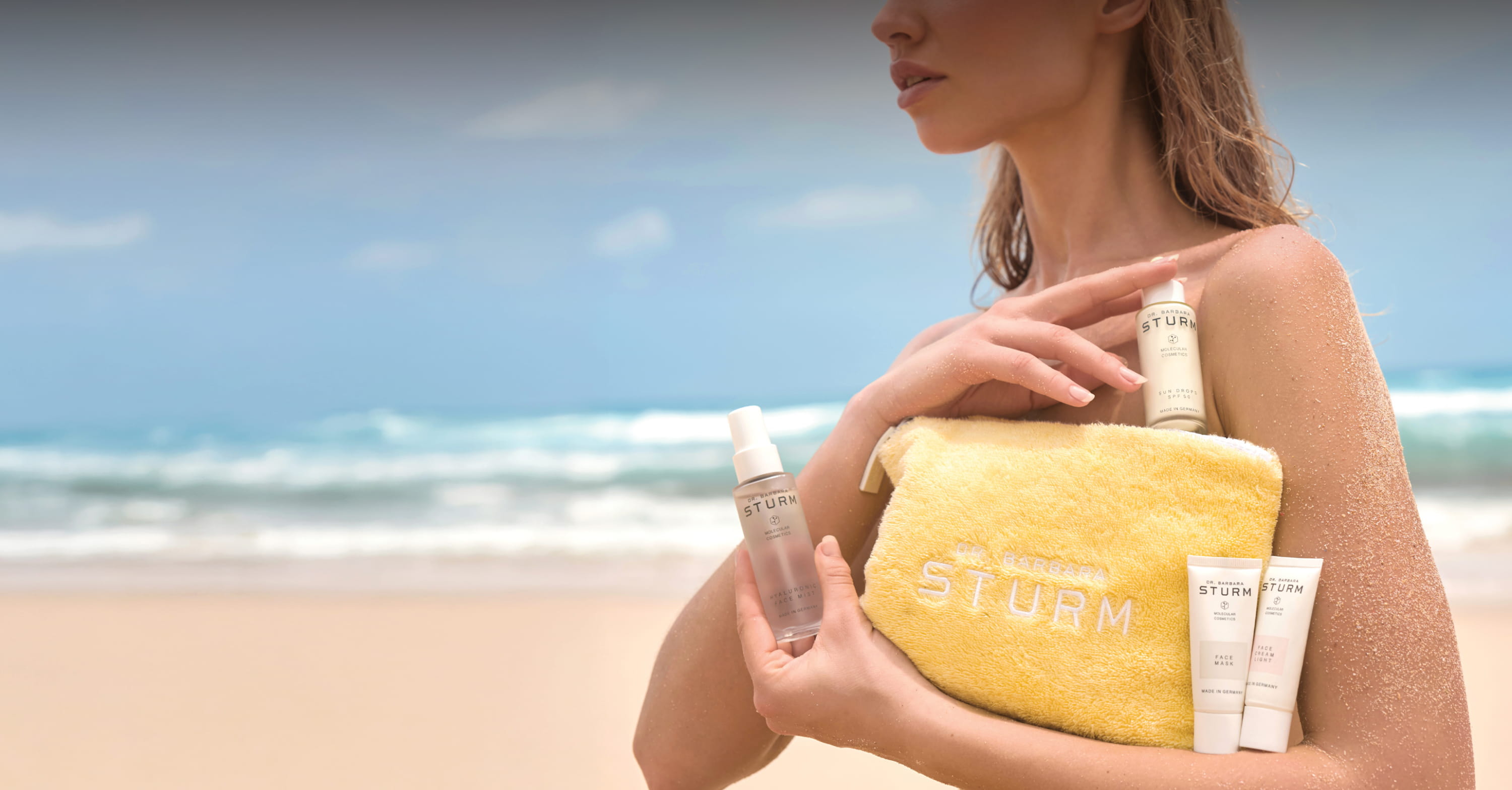 Woman holding a yellow pouch with skincare products on a beach.