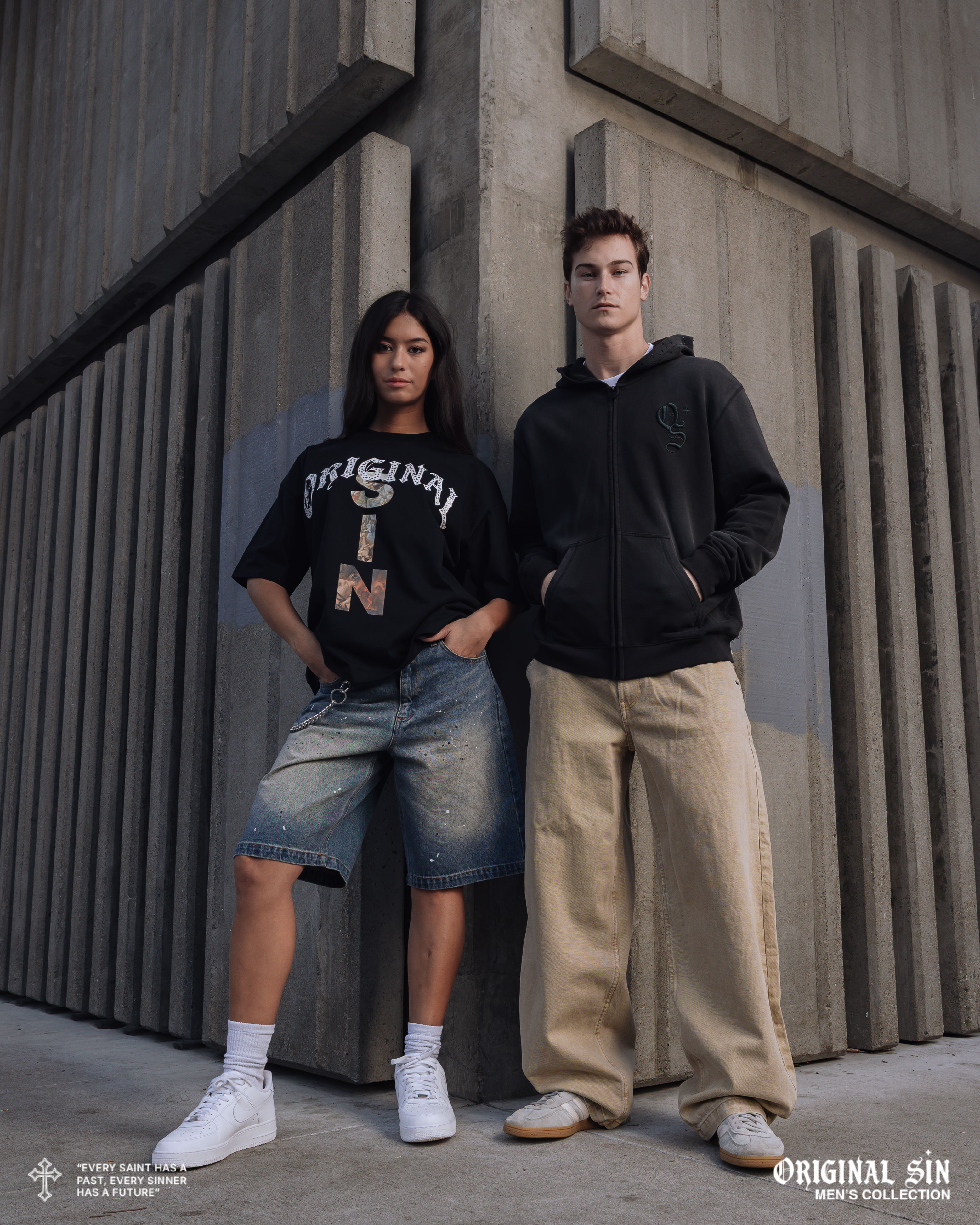 Two young adults posing in streetwear against a concrete wall.