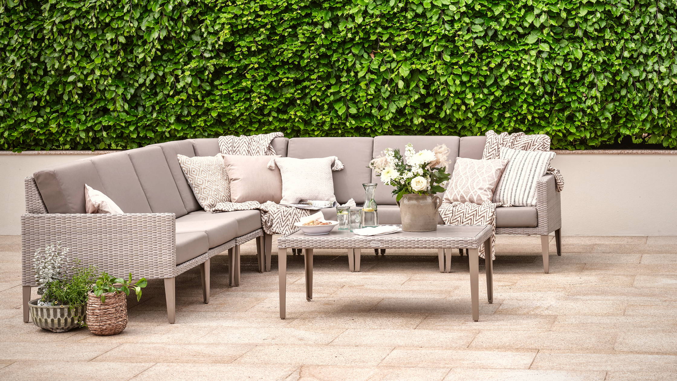 Rattan garden furniture modular sofa set covered with beige cushions in a hedged garden.