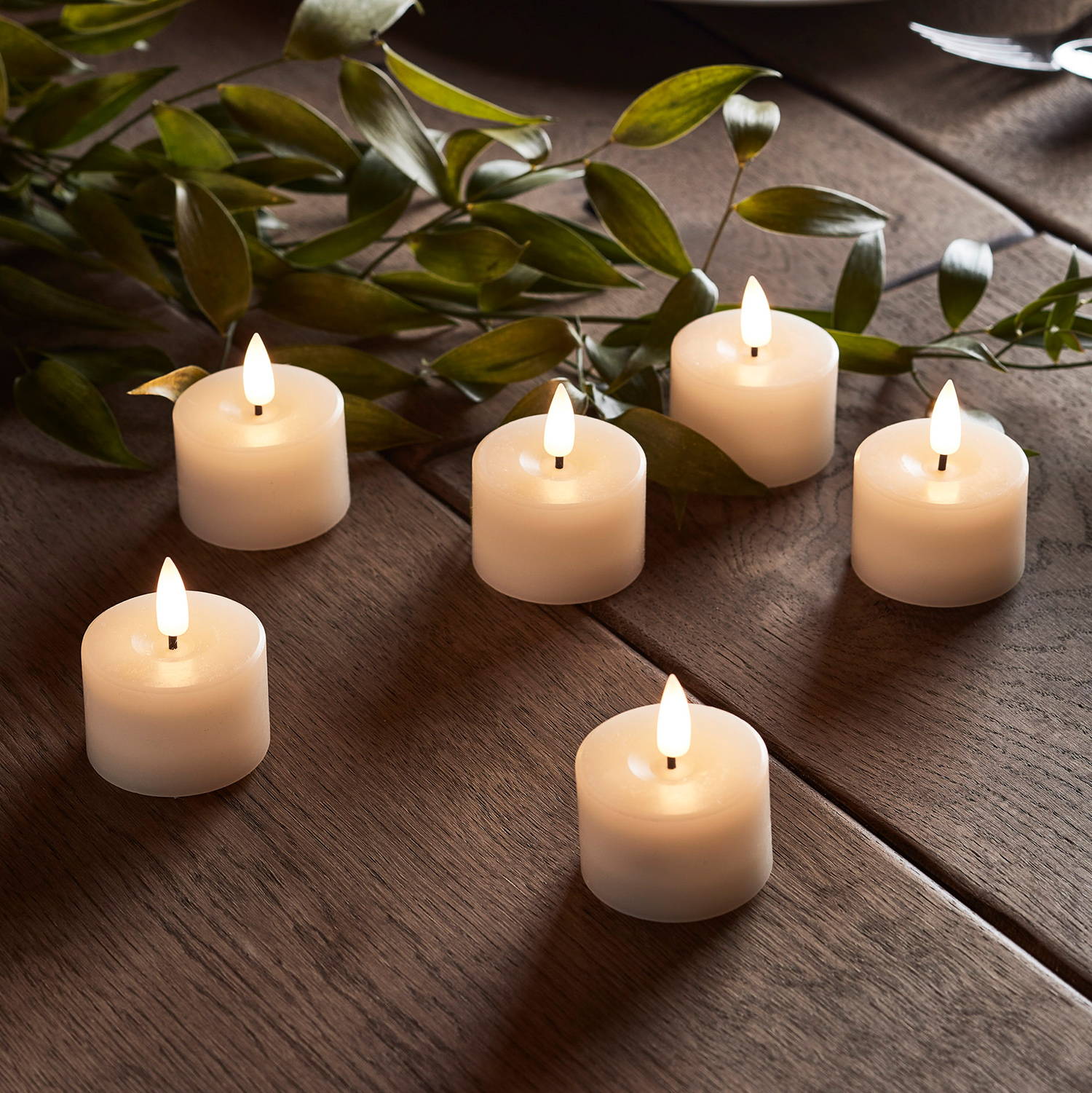 6 mini ivory votive LED candles on a wooden floor with some faux foliage.